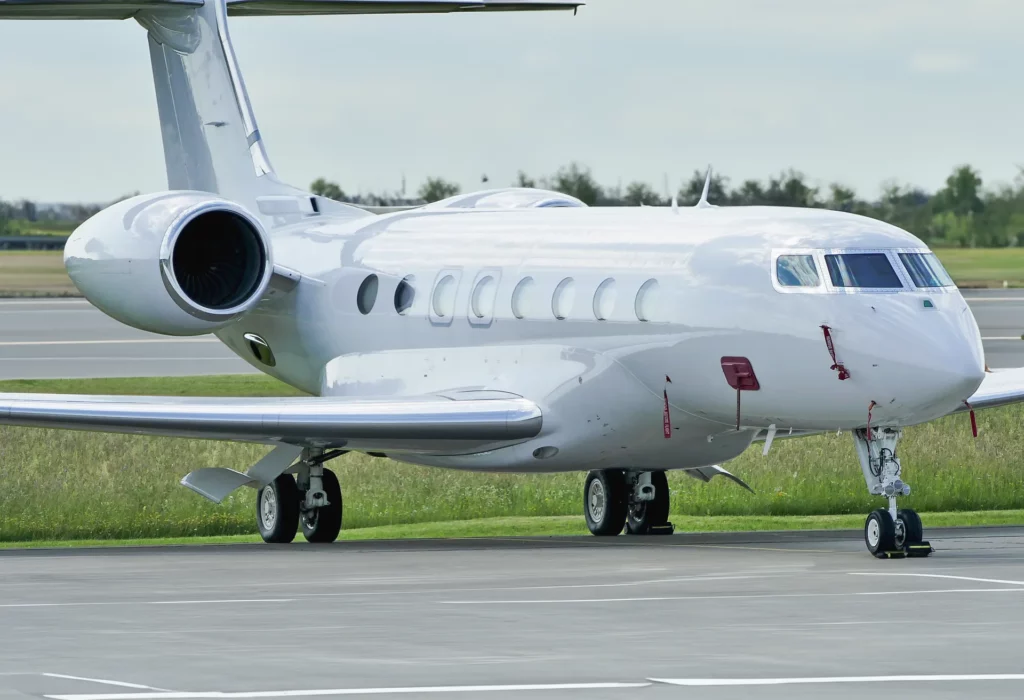 A white Gulfstream G650 private jet is parked on a runway, with its sleek, modern design and multiple windows visible. The jet's landing gear is down, and its powerful engine can be seen on the left side of the aircraft. Trees and grass are in the background, indicating it is at an airport.