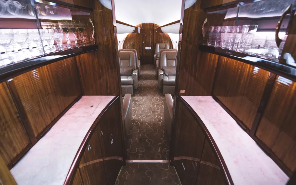 A luxurious galley inside the Bombardier Global 5000 featuring glossy wood paneling, a marble-like countertop, and glassware neatly displayed on shelves. The aisle leads toward plush seating, with ambient lighting creating an elegant atmosphere.