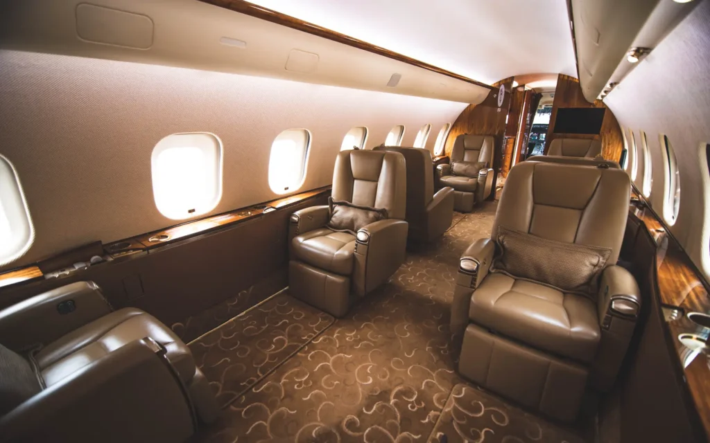 The interior of the Bombardier Global 5000 featuring spacious executive-style seating with wooden tables. The neutral-toned leather chairs provide ample legroom, and a flat-screen monitor is mounted on the side panel for in-flight entertainment.