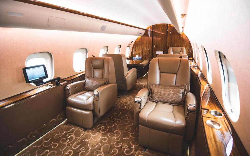 A view inside the private jet’s forward cabin, showing pairs of reclining leather seats arranged around glossy wooden tables. The soft lighting, multiple windows, and elegant decor create a refined travel experience.