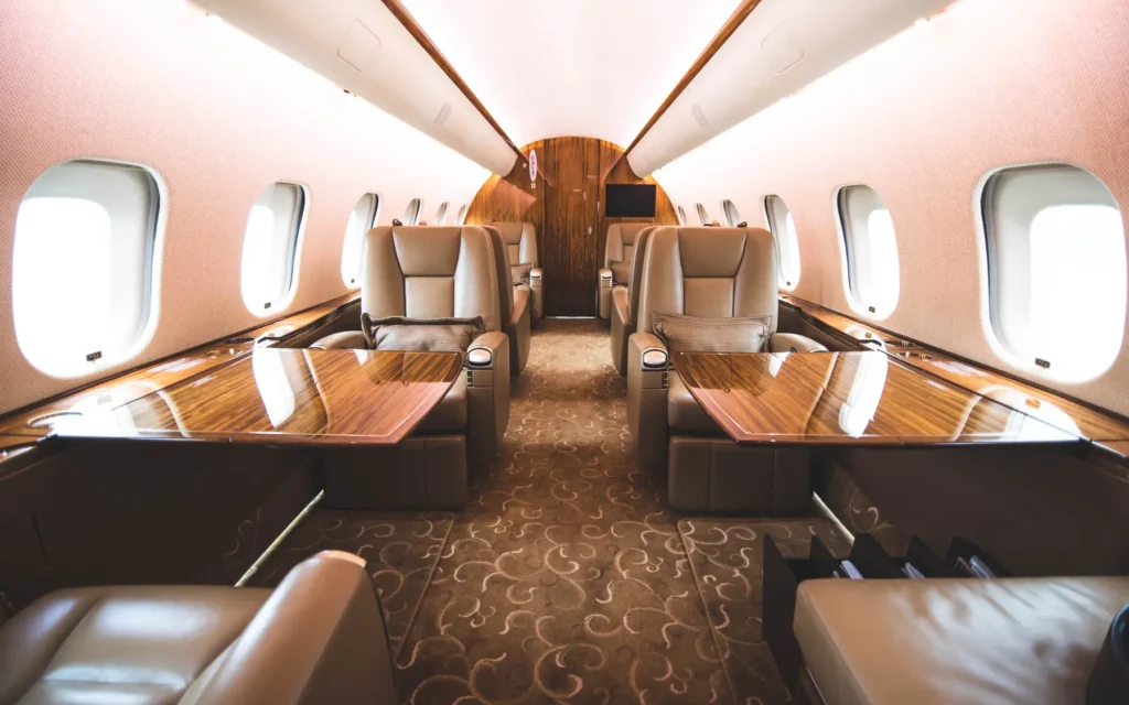 A row of plush, reclining leather seats with armrests and accent pillows, arranged alongside the windows in the private jet’s cabin. The wood-paneled walls and carpeted flooring add to the luxurious setting, with a partition leading toward the cockpit area.
