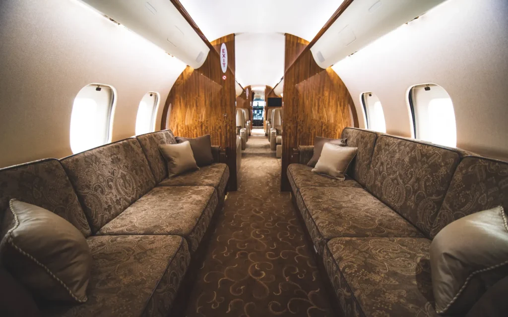 A spacious lounge area inside the private jet, featuring two long, fabric-upholstered divans with decorative pillows, positioned along the windows. The warm-toned wooden accents, soft lighting, and plush carpeting enhance the cozy and sophisticated ambiance.