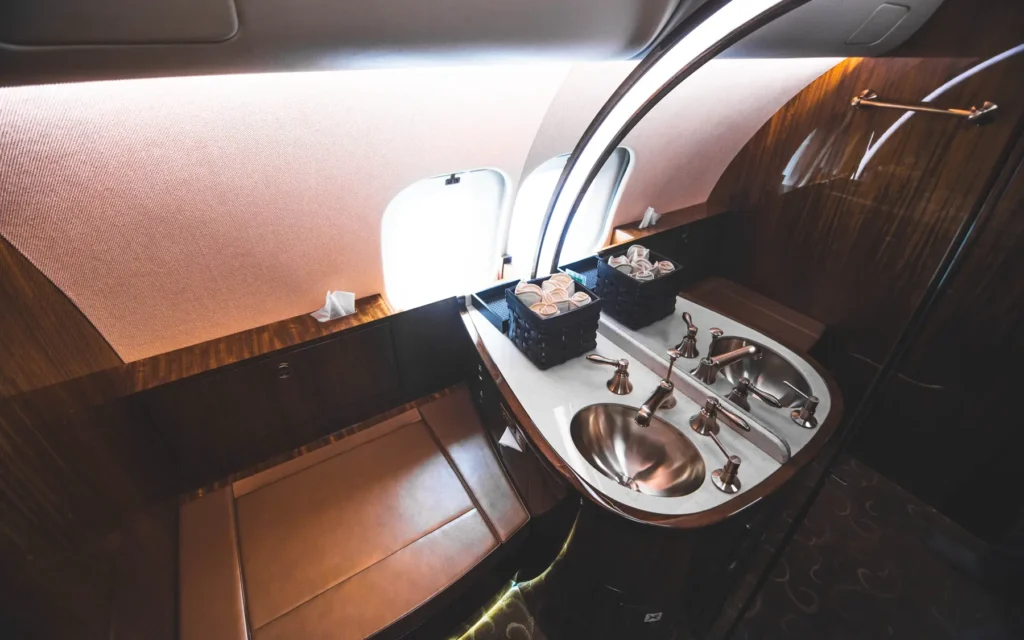 A luxurious private jet lavatory with dual stainless steel sinks, polished faucets, and a marble-style countertop. A cushioned bench seat sits beneath the windows, while neatly organized amenities complete the refined space.