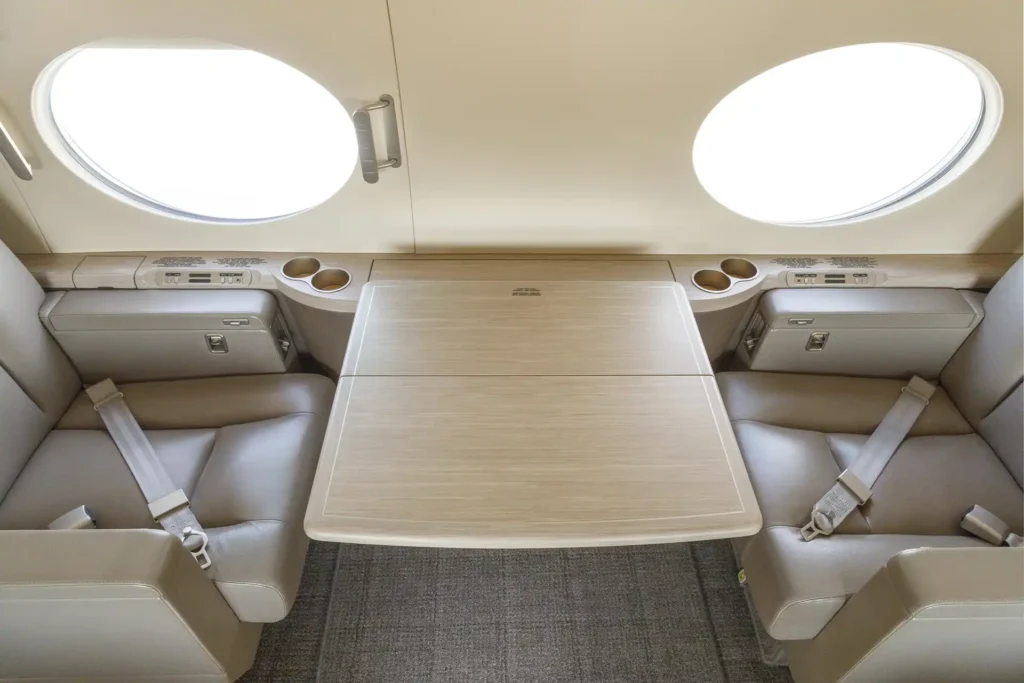 Overhead view of four cabin seats arranged around a large wooden fold-out table, inside Gulfstream G550 SN:5562, with visible seatbelts and dual window views on each side.