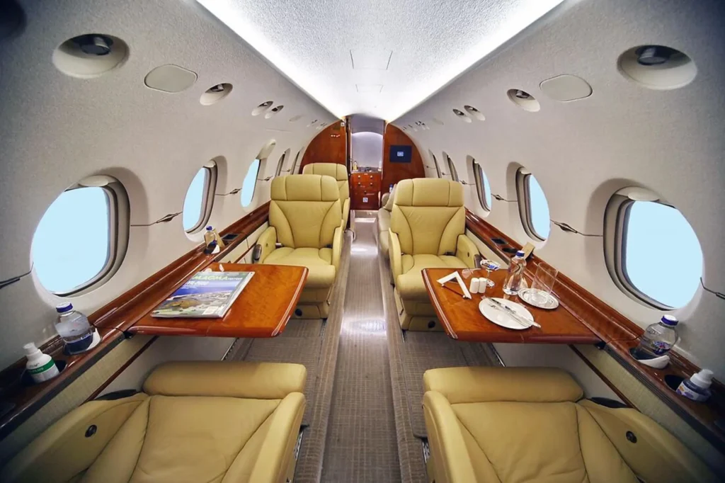 Wide view down a private jet cabin with tan leather seats and glossy wood side tables beside oval windows. Bottled water and a magazine sit on the tables under soft overhead lighting.