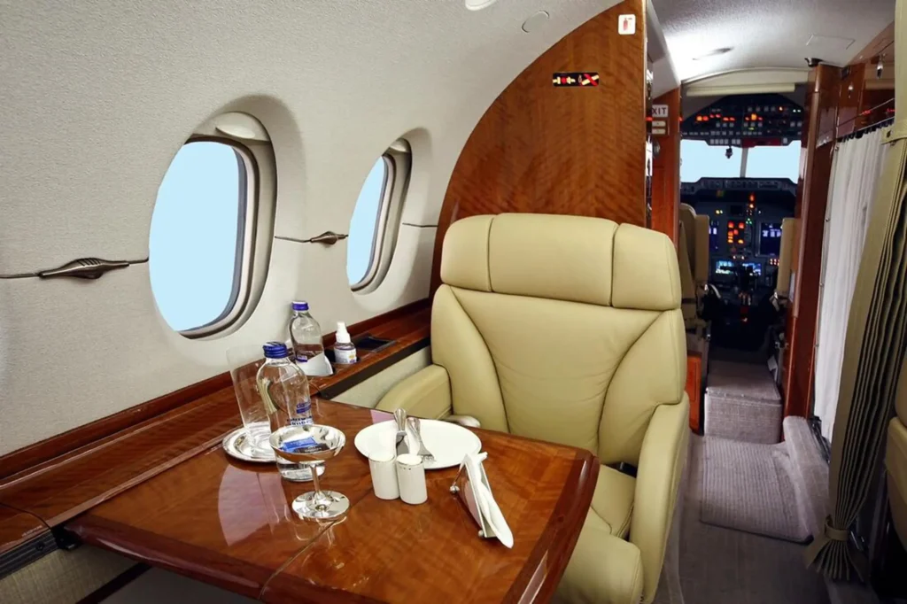 Close view of a single tan leather seat and wood table set with glassware and a folded napkin inside a private jet. The cockpit is visible in the background through the aisle.