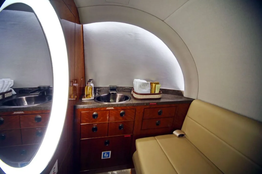 Private jet lavatory area with a curved wall, oval mirror, sink, and wood cabinetry. A tan bench seat sits beside the counter with toiletries arranged near the sink.