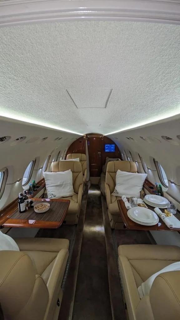 Private jet cabin set for dining with tan leather seats, wood tables, and white plates and napkins. Pillows embroidered “Dio” rest on the seats and the aisle leads toward the front bulkhead.