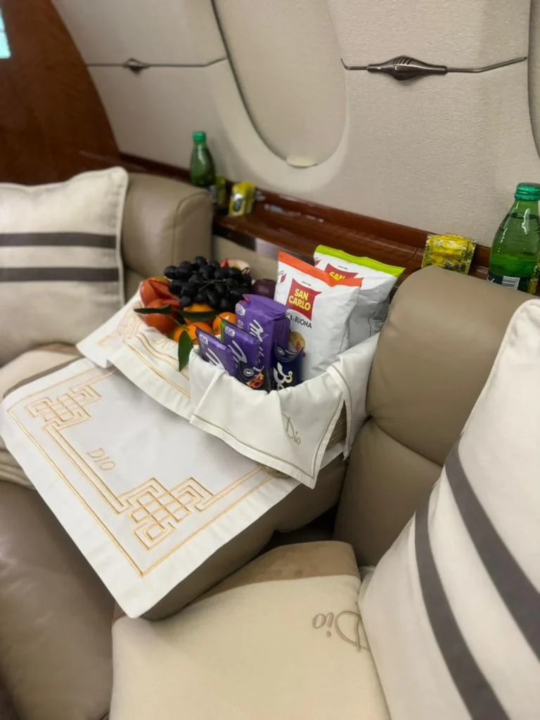 Snack setup on a private jet seat with a white cloth embroidered “Dio” and a basket of treats. Visible labels include San Carlo chips and wrapped snacks with green glass bottles in the background.