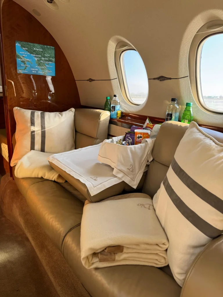 Lounge area on a private jet with tan leather couch, striped pillows, and folded blanket. A snack basket sits on a white cloth embroidered “Dio” near the windows with a small map screen on the wood panel.