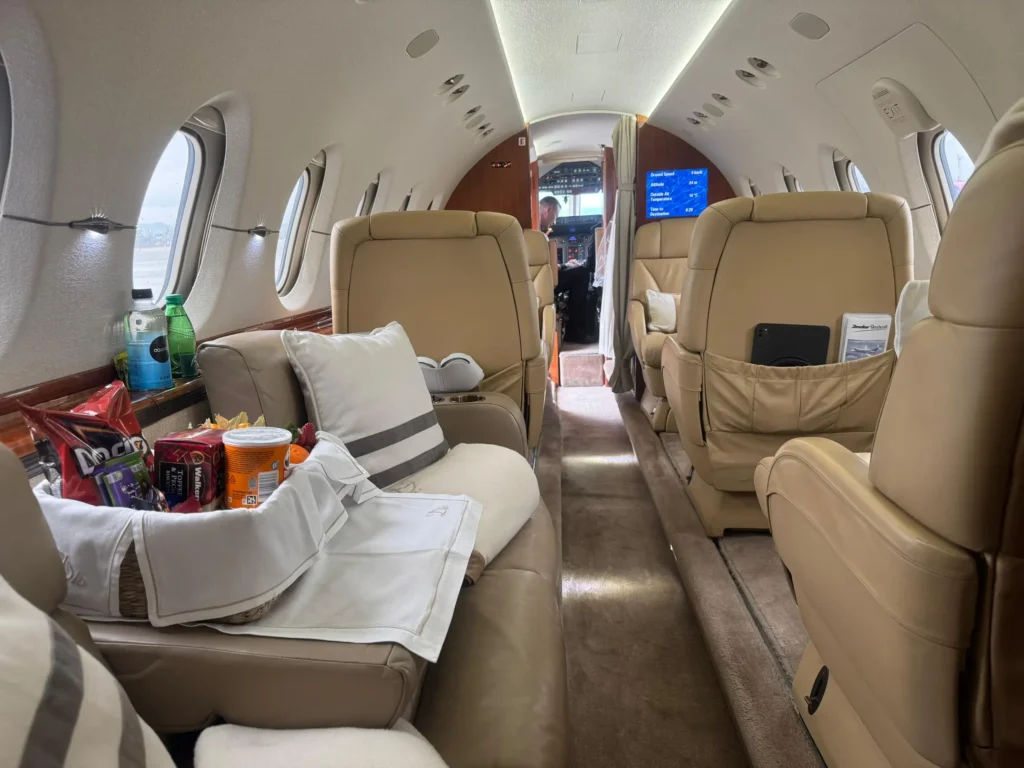 View down a private jet cabin with tan leather seats and a snack basket on a side couch. A screen near the cockpit shows flight information while drinks and snacks sit along the window ledge.